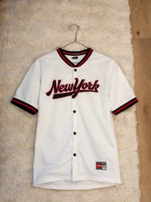 Vit baseballskjorta med röd text - Säljer en vit baseballskjorta med knappar och röd-svart text på bröstet. Skjortan har svarta och röda detaljer på krage och ärmslut, och är i en loose fit. Perfekt för dig som gillar sportig och avslappnad stil. Storlek S I herr modell 