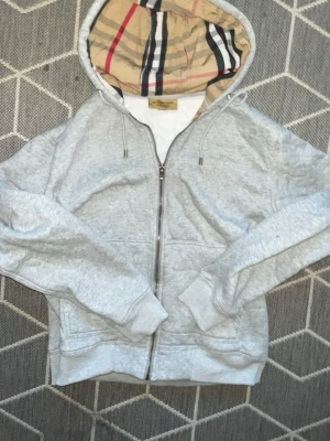 Grå hoodie från Burberry med huva - Snygg grå zip-hoodie från Burberry med den klassiska beigea, röda och svarta rutan i huvan. Hoodien har dragsko i huvan, två fickor fram och ribbade muddar. Perfekt för dig som vill ha en stilren men ändå lyxig look. Materialet känns mjukt och skönt.