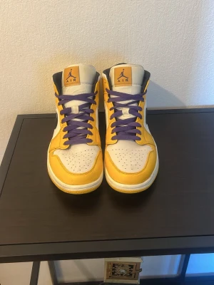 Air Jordan 1 High gul/vit/lila - Säljer ett par Air Jordan 1 High sneakers i gult och vitt med lila skosnören och svart swoosh. Klassisk high-top siluett med perforerad tåbox och ikonisk Jumpman-logga på plösen. Ovandelen är i läder och sulan är vit med gula detaljer.