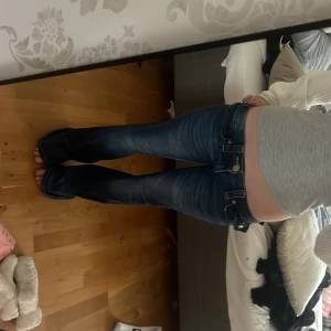 Blå bootcut jeans från True Religion - Säljer ett par blå bootcut jeans från True Religion med snygga slitningar och kontrastsömmar. Jeansen har låg midja, fem fickor och detaljerade bakfickor med broderi och knappar. Perfekta för en avslappnad men trendig look.