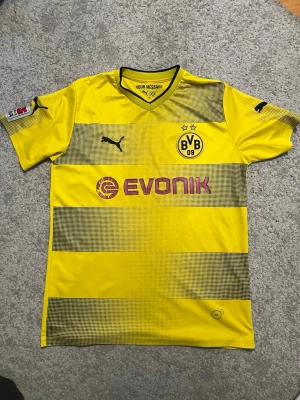 Gul Borussia Dortmund matchtröja Puma - Säljer en gul Borussia Dortmund matchtröja från Puma med svarta detaljer och tryck. Tröjan har klubbmärke, sponsorlogga och nummer 17 Aubameyang på ryggen. Korta ärmar, V-ringning och tillverkad i lätt funktionsmaterial.