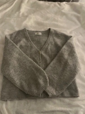 Ljusgrå kofta med v-ringning - Mysig ljusgrå kofta med v-ringning och fyra knappar framtill. Koftan har långa ärmar och är stickad i ett mjukt material, perfekt för lager på lager. Enkel och stilren design som passar till det mesta.