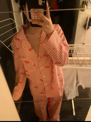 Rödvit randig pyjamas med rosetter - Säljer en söt pyjamas med rödvita vertikala ränder och små röda rosetter som detalj. Setet har klassisk krage, knappstängning framtill och en bröstficka. Perfekt för mysiga kvällar hemma.