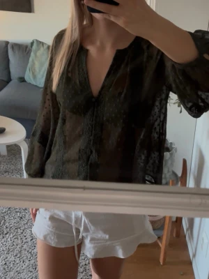 Transparent svart blus med prickar - Snygg transparent svart blus med diskreta prickar och v-ringning. Blusen har långa ärmar och ett lätt, luftigt material som ger en avslappnad men trendig look. Perfekt att styla med ett linne under för en cool layering-effekt.