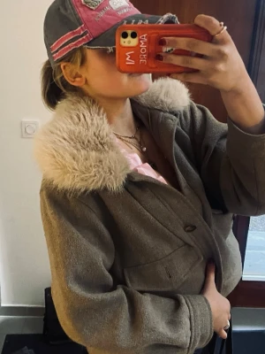 Kort beige kappa med fuskpäls - Snygg beige kappa i ullmix med stor fluffig fuskpäls-krage. Modellen är kort och boxig med stora fickor framtill och knappar. Perfekt för dig som vill ha en trendig och mysig look i höst och vinter.