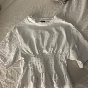 Populär Gina ribbad tröja - Säljer en vit t-shirt med korta ärmar och insydd midja som ger en snygg siluett. Materialet är mjukt och ribbat, perfekt för dig som gillar stilrena plagg med lite extra detalj. Passformen är normal och t-shirten har en klassisk rund hals.