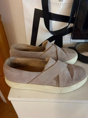 Beige slip-on sneakers i mocka - Snygga beige slip-on sneakers med korslagda band över foten. Tillverkade i mjuk mocka med vit sula och perforerad innersula för extra komfort. Perfekta för en avslappnad och trendig stil. Köpta på sellpy men originalmärket är zign. 