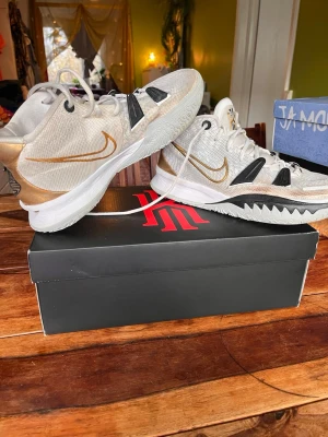 Nike Kyrie vita och guld sneakers - Snygga Nike Kyrie sneakers i vitt med gulddetaljer och svarta inslag. Skorna har mesh-överdel för bra andningsförmåga, gummisula och coola detaljer på sidan. Perfekta för dig som gillar basketstil och vill sticka ut med unika färgkombos. Och vill ge dig själv den lilla extra boosten på basket planen 