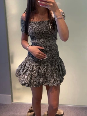 Svart blommig offshoulder klänning - Söt svart kortklänning med vita små blommor och offshoulder-design. Klänningen har volang nertill och smockad överdel som ger en feminin siluett. Perfekt för varma dagar och sommarens alla tillfällen.