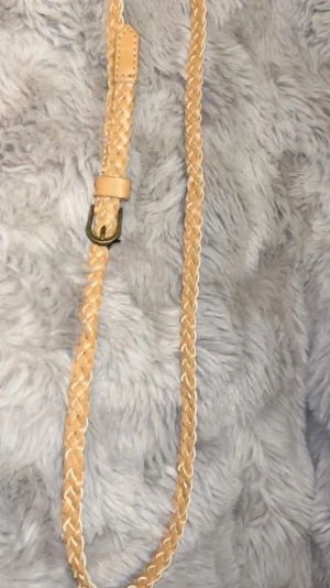 Flätat beige bälte i skinn - Snyggt flätat bälte i beige skinn med metallspänne. Perfekt för att ge din outfit en stilren touch. Bältet har en klassisk design och passar till många olika looks.