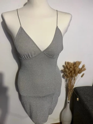 Glittrig silver kortklänning - Snygg glittrig silverfärgad kortklänning med tunna axelband och v-ringad urringning. Klänningen har en figurnära passform och är perfekt för dig som vill sticka ut. Materialet känns mjukt och stretchigt, vilket gör den bekväm att bära. Axelbandet är sydda lite för att göra klänningen mindre. Man kan klippa bort det så kommer dem vara som förr 