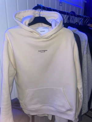 Beige hoodie från Axel Arigato - Snygg beige hoodie från Axel Arigato med minimalistisk logga på bröstet. Hoodien har en klassisk känguruficka och mjuk insida. Perfekt för dig som gillar stilrena plagg med en modern touch. Tillverkad i bomullsmix för skön komfort.
