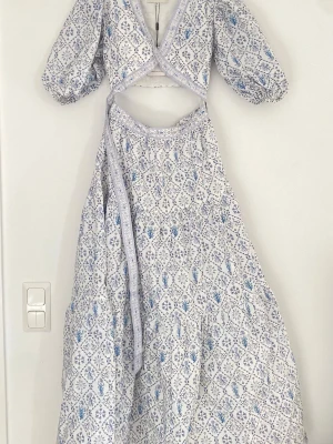 Vit blommig omlottklänning By Malina - Vit omlottklänning från By Malina med lila och blått blommönster. Klänningen har puffiga trekvartsärmar, v-ringning och volangdetaljer längs kanterna. Tillverkad i bomull och perfekt för sommarens alla tillfällen.