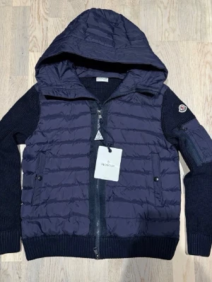 Mörkblå dunjacka från Moncler - Snygg mörkblå dunjacka från Moncler med stickade ärmar och huva. Jackan har dragkedja framtill, två fickor med knappar och en Moncler-logga på ärmen. Perfekt för kalla vinterdagar och riktigt trendig look.