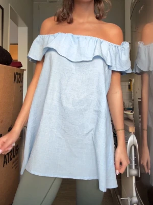 Ljusblå offshoulder klänning med volang - Söt ljusblå kortklänning i bomull med bred volangkant upptill. Klänningen har offshoulder-design och luftig passform, perfekt för varma dagar. Kan även bäras oneshoulder för en trendig look.
