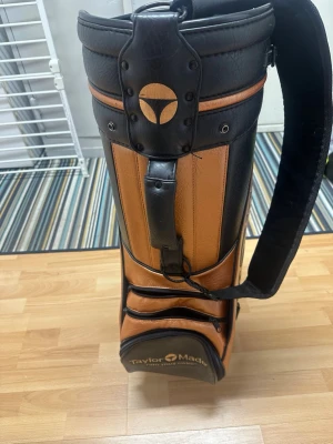 TaylorMade golfbag i brunt och svart - Snygg golfbag från TaylorMade i brunt och svart konstläder med flera fack och axelrem. Klassisk design med tydlig logga på sidan och detaljer i svart. Perfekt för att bära golfklubbor och tillbehör på banan.