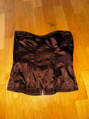 Brun satin korsettopp - Snygg brun korsettopp i satin med markerade sömmar.  Toppen är axelbandslös och har dragkedja bak för enkel påklädning. Perfekt för att lyfta vilken outfit som helst. Inget tjockt tyg. 