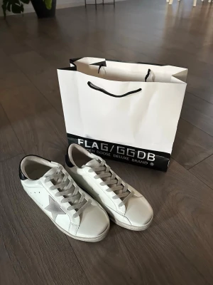 Golden Goose vita sneakers med stjärna - Säljer ett par vita Golden Goose sneakers med grå stjärna på sidan, grå skosnören och svart detalj på hälen. Klassisk låg modell i läder med slitna detaljer för en cool och avslappnad look. Perfekta för dig som gillar streetstyle och trendiga skor.