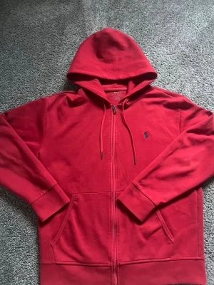 Röd hoodie från Polo Ralph Lauren - Röd hoodie med dragkedja från Polo Ralph Lauren. Klassisk design med huva och dragsnören, två fickor framtill och den ikoniska blå broderade loggan på bröstet. Tillverkad i mjukt bomullsmaterial som är skönt att ha på sig.