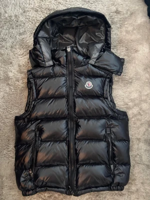 Svart dunväst från Moncler med huva - Snygg svart dunväst från Moncler med avtagbar huva och två dragkedjefickor framtill. Västen har en glansig finish, Moncler-logga på bröstet och breda quiltade paneler. Perfekt för lager-på-lager och streetstyle.