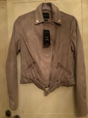 Beige skinnjacka från New Look - Snygg beige skinnjacka från New Look med asymmetrisk dragkedja, två snedställda fickor med dragkedja och bälte i midjan. Klassisk bikerstil med krage och tryckknappar. Perfekt för en trendig look.