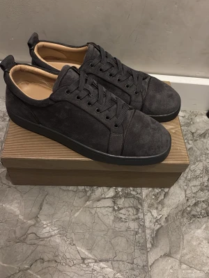 Mörkgråa sneakers från Christian Louboutin - Snygga mörkgråa sneakers i mocka från Christian Louboutin med klassisk röd sula. Skorna har lågt skaft, svarta skosnören och en stilren design. Perfekta för dig som vill ha något exklusivt och trendigt i garderoben.