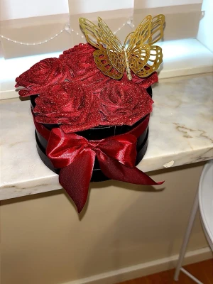 Evighetsrosor röda Glitter rosor Love - Säljer en rund svart dekorationsask fylld med glittriga röda konstrosor och en stor röd sidenrosett. På toppen sitter en guldfärgad metallfjäril som ger en lyxig känsla. Perfekt som present eller inredningsdetalj.