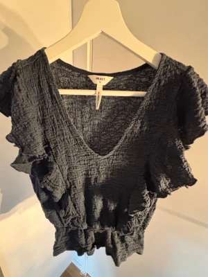 Mörkgrå volangtopp från Object - Supersnygg mörkgrå topp från Object med volangdetaljer längs ärmarna och v-ringad hals. Tillverkad i ett krinklat, lätt material som ger en avslappnad men trendig look. Perfekt att styla med jeans eller kjol för en cool vibe.