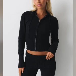 Svart stickad hoodie sett - Svart stickad hoodie från Gina Tricot med croppad passform, ribbade detaljer i midjan och på ärmsluten. Jackan har dragkedja framtill, huva med snörning och långa ärmar. Perfekt för en chill och trendig look.