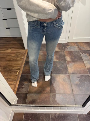 Blå bootcut jeans från True Religion - Säljer ett par ljusblå bootcut jeans från True Religion med slitningar och snygga detaljer på bakfickorna. Jeansen har låg midja, klassisk femficksmodell och är tillverkade i denim med stretch för skön passform. Byxorna är endast använde ett fåtal gånger och har bara lite slitning nere vide foten 