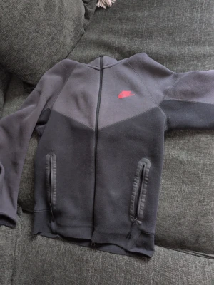 Nike tech fleece - Snygg svart och grå hoodie från Nike med dragkedja framtill och två fickor med dragkedja. Ikonisk Nike-logga i rött på bröstet. Hoodien har en sportig look och är tillverkad i mjukt material som passar perfekt till streetwear. Storlek S. Säljer pga för liten