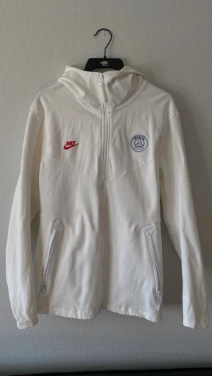 Vit Nike PSG hoodie - Snygg vit hoodie från Nike med PSG-logga på bröstet och röd Nike Swoosh. Hoodien har dragkedja framtill, två fickor med dragkedja och en stor huva. Tillverkad i mjukt material som känns skönt mot huden. Aldrig använd