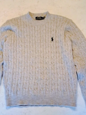  kabelstickad tröja Polo Ralph Lauren - Kabelstickad beige tröja från Polo Ralph Lauren med klassisk svart broderad logga på bröstet. Tröjan har rund halsringning, ribbade muddar och är tillverkad i mjukt material som känns lyxigt och bekvämt. Perfekt för dig som gillar stilrena och tidlösa plagg.