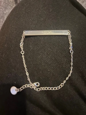 Silverfärgat armband från Wera - Stilrent armband i silverfärg från Wera med en rektangulär platta och tunn kedja. Plattan har ingraverad logga och armbandet har justerbar längd med en liten rund bricka vid låset. Perfekt accessoar för en clean look.
