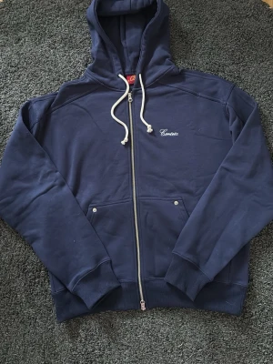 Mörkblå Corteiz hoodie  - Mörkblå hoodie från Corteiz dragkedja framtill, vita dragsnören i huvan och två fickor med knappdetaljer. Broderad logga på bröstet och ribbade muddar vid ärmar och nederkant. Perfekt för en avslappnad stil. Pris kan diskuteras!