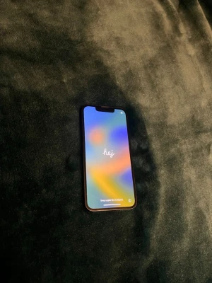 Svart iPhone 13 mini 256gb (100% batterikapacitet) - Snygg svart iPhone med stilren design och dubbla kameror på baksidan. Telefonen har en stor skärm med tunn ram och glasbaksida med Apple-loggan. Perfekt för dig som gillar modern teknik och vill ha en mobil som känns både lyxig och smidig.