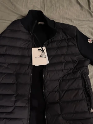 Mörkblå cardigan från Moncler - Snygg mörkblå cardigan från Moncler med quiltad design och dragkedja framtill. Västen har en hög krage och Moncler-logga på ärmen. Tillverkad i ett glansigt material med dunfyllning för extra värme. Perfekt för lager-på-lager och streetstyle.