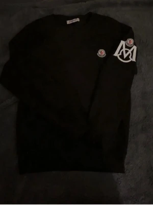 Svart sweatshirt från Moncler - Svart sweatshirt från Moncler med klassisk logotyp på bröstet och stort vitt M-tryck på ena ärmen. Tröjan har rund halsringning och är tillverkad i mjukt bomullsmaterial. Perfekt för dig som gillar streetwear och exklusiva märken.