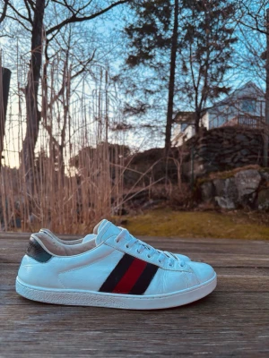 Gucci ace - Snygga vita sneakers från Gucci i läder med klassiska röda och mörkblå ränder på sidan. Skorna har lågt skaft, vita snören och en svart detalj på hälen. Perfekta för dig som vill ha en clean och stilren look med lyxig känsla.
