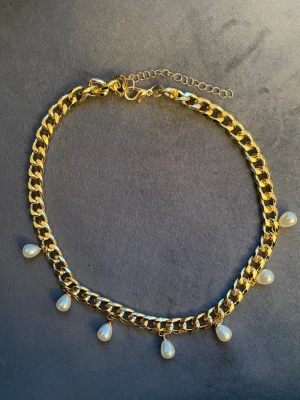 Guldfärgat halsband med pärldroppar - Snyggt halsband i guldfärgad metall med bred kedja och sex droppformade vita pärlor som hänger längs kedjan. Justerbar längd med förlängningskedja. Perfekt accessoar för att lyfta din outfit.