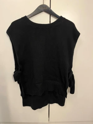 Svart stickad väst med knytband - Snygg svart stickad väst med ribbade kanter och dekorativa knytband i sidorna. Perfekt att bära över en skjorta eller t-shirt för en trendig lager-på-lager-look. Rund halsringning och avslappnad passform.