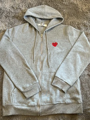 Grå hoodie från Comme des Garçons Play - Snygg grå zip-hoodie från Comme des Garçons Play med det ikoniska röda hjärtat med ögon broderat på bröstet. Hoodien har dragkedja, två fickor fram och ribbade muddar. Tillverkad i mjuk bomullsblandning och har en klassisk huva med dragsko.