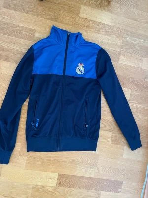 Blå Real Madrid träningsjacka - Snygg träningsjacka i marinblått och blått från Real Madrid. Jackan har dragkedja framtill, två fickor med dragkedja och klubbens logga på bröstet. Tillverkad i ett mjukt och stretchigt material, perfekt för sport eller chill.