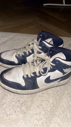 Air Jordan 1 Mid amory navy sneakers - Säljer ett par Air Jordan 1 Mid sneakers i blått och vitt läder med klassisk Nike Swoosh och Air Jordan-logga på sidan. Skorna har vita snören och en vadderad krage för extra komfort. Perfekta för dig som gillar streetwear och ikonisk design. Pris går även att förhandla :)