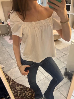 Vit offshoulder topp i bomull - Söt vit offshoulder topp med korta puffärmar och rak, lös passform. Tillverkad i mjuk bomull och har en smockad kant upptill som ger en trendig och avslappnad look. Perfekt att styla med jeans eller kjol.
