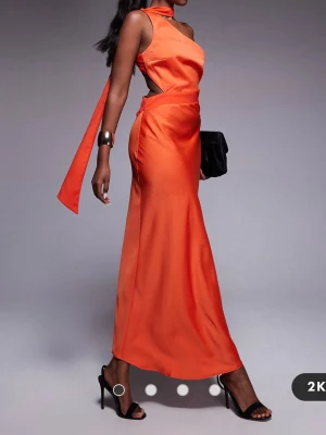 Orange långklänning med öppen rygg - Snygg orange långklänning i satin med oneshoulder och öppen rygg. Klänningen har en elegant slits och ett brett band som knyts i ryggen. Perfekt för dig som vill sticka ut med en färgstark look.