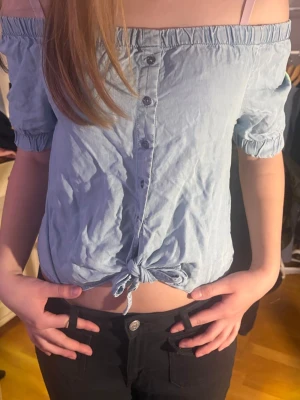 Ljusblå offshoulder knytblus - Trendig ljusblå blus med offshoulder och korta ärmar. Knappar framtill och knytning i midjan ger en avslappnad och snygg look. Perfekt till jeans eller kjol för en cool stil.