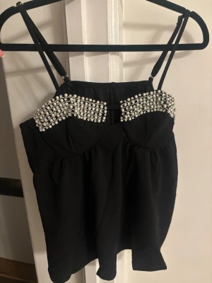 Svart korsettopp med pärlor - Snygg svart korsettopp med dubbla smala axelband och dekorerad med pärlor över bysten. Toppen har en figurnära passform upptill och ett lätt utställd fall nedtill. Perfekt för dig som vill sticka ut med en unik detalj.
