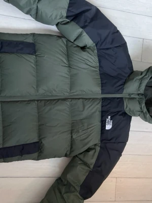 Grön och svart dunjacka The North Face - Säljer en grön och svart dunjacka från The North Face med huva och dragkedja. Jackan har stora, quiltade partier och logga på bröstet och ryggen. Perfekt för kalla vinterdagar och riktigt snygg streetstil.