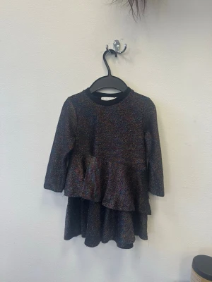 Glittrig svart volangklänning H&M - En svart glittrig klänning från H&M med volangdetaljer och lång ärm. Klänningen har en rund halsringning och skimrar i flera färger. Perfekt för dig som vill sticka ut lite extra.